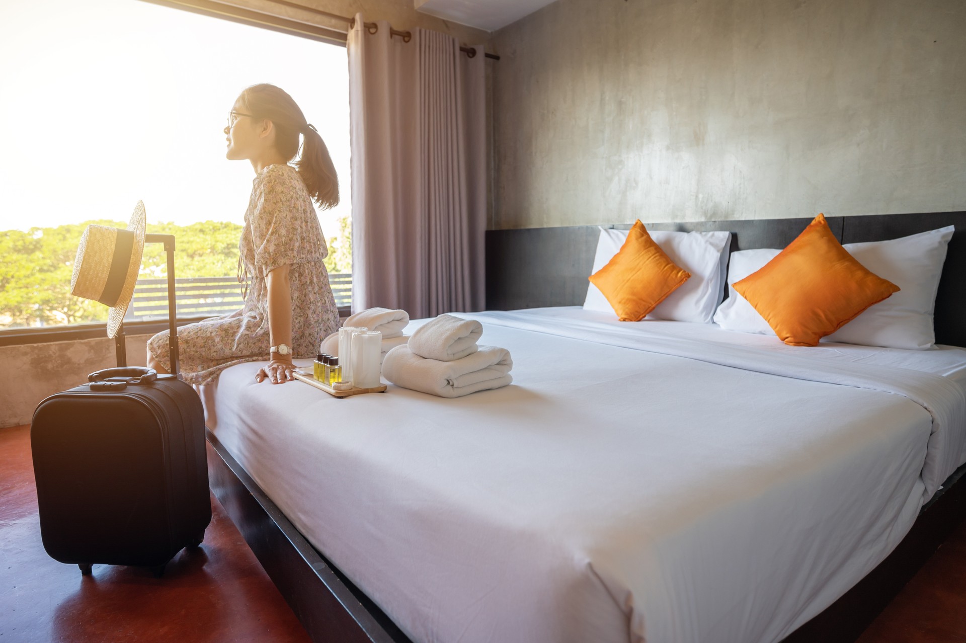 Tourist woman sitting on bed nearly window, looking to beautiful view with her luggage in hotel bedroom after check-in.